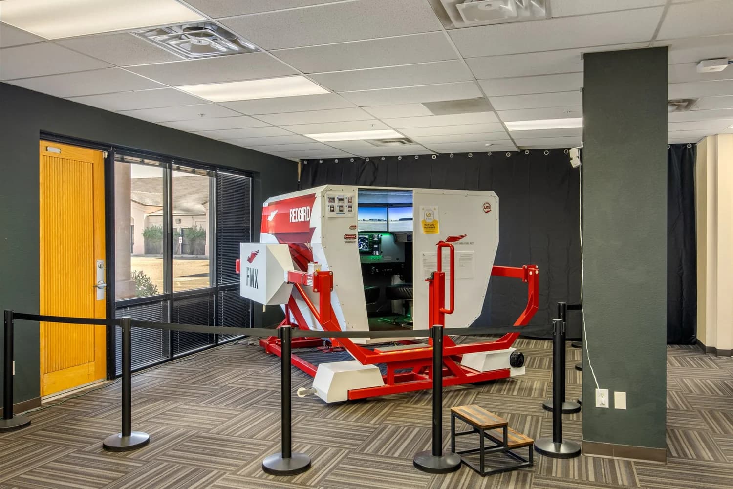 Redbird FMX advanced aviation training device — a red and white full-motion enclosed cab simulator on its motion base, behind crowd-control stanchions