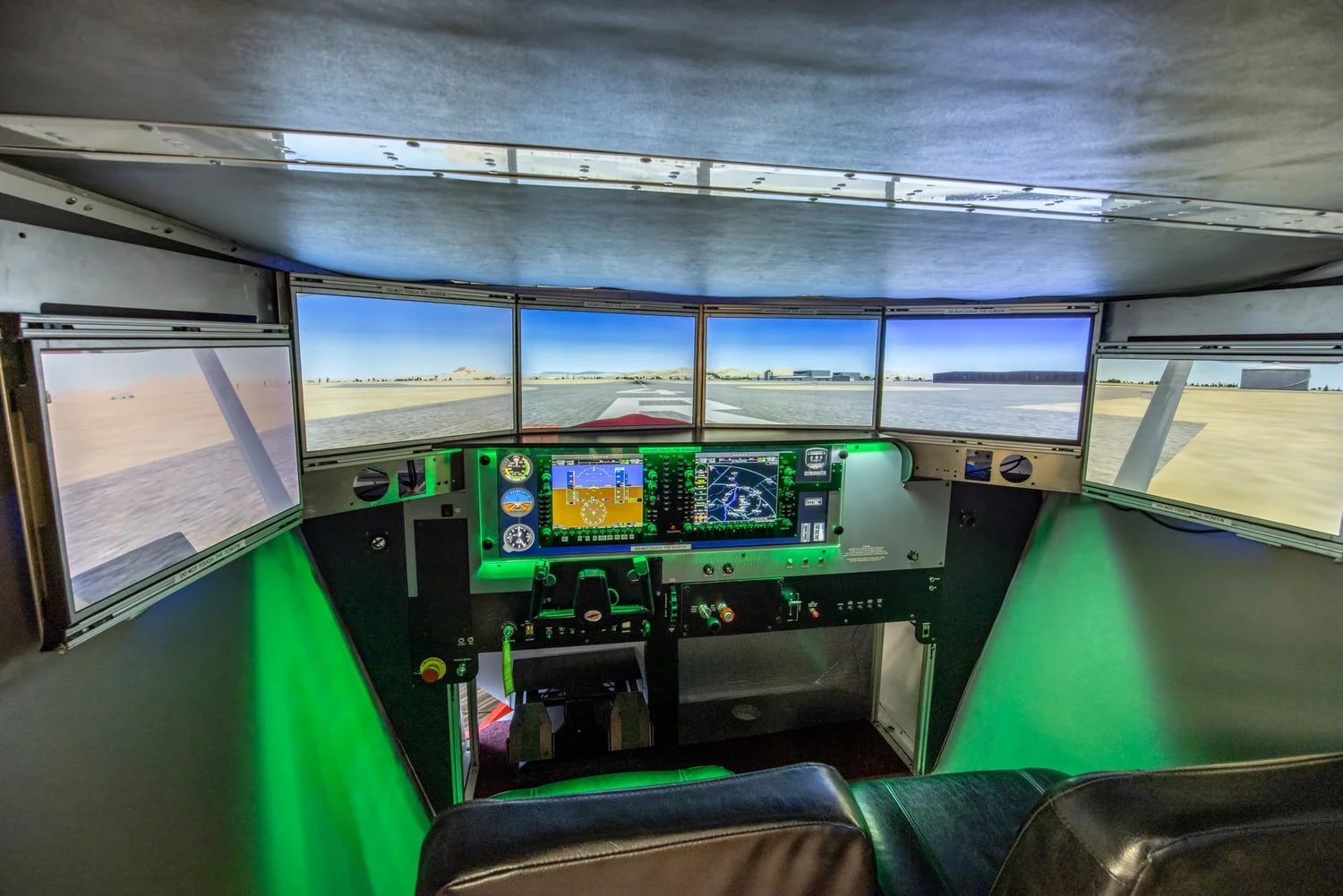 Inside the Redbird FMX cockpit at night — six wraparound visual screens showing a runway, twin glass-cockpit displays, dual yokes, and atmospheric green lighting