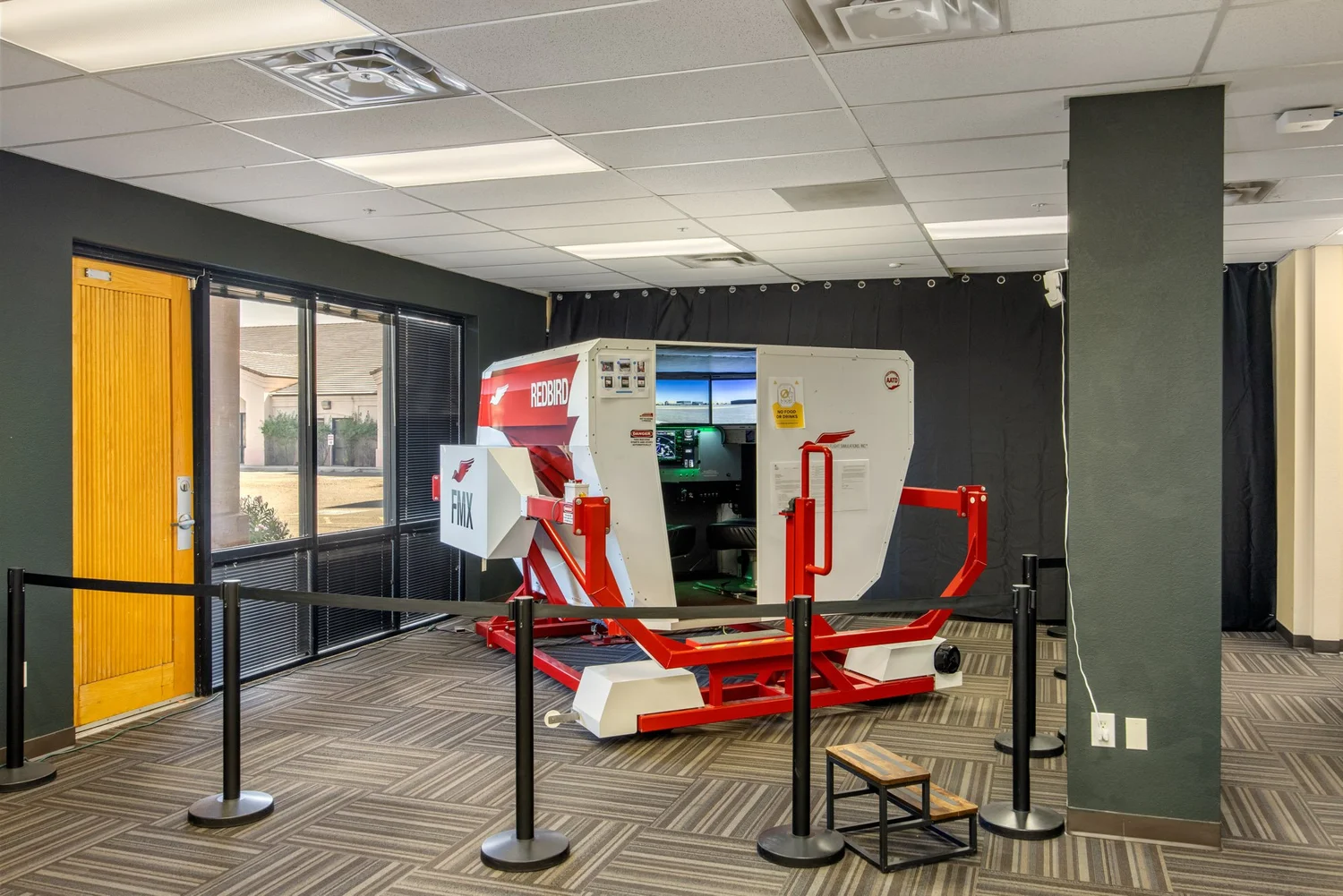 Redbird FMX advanced aviation training device — a red and white full-motion enclosed cab simulator on its motion base, behind crowd-control stanchions