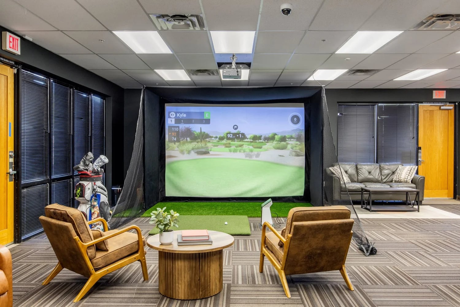 Tour-grade golf simulator bay mid-round, projector throwing a fairway across a 16-foot screen with leather lounge chairs in front